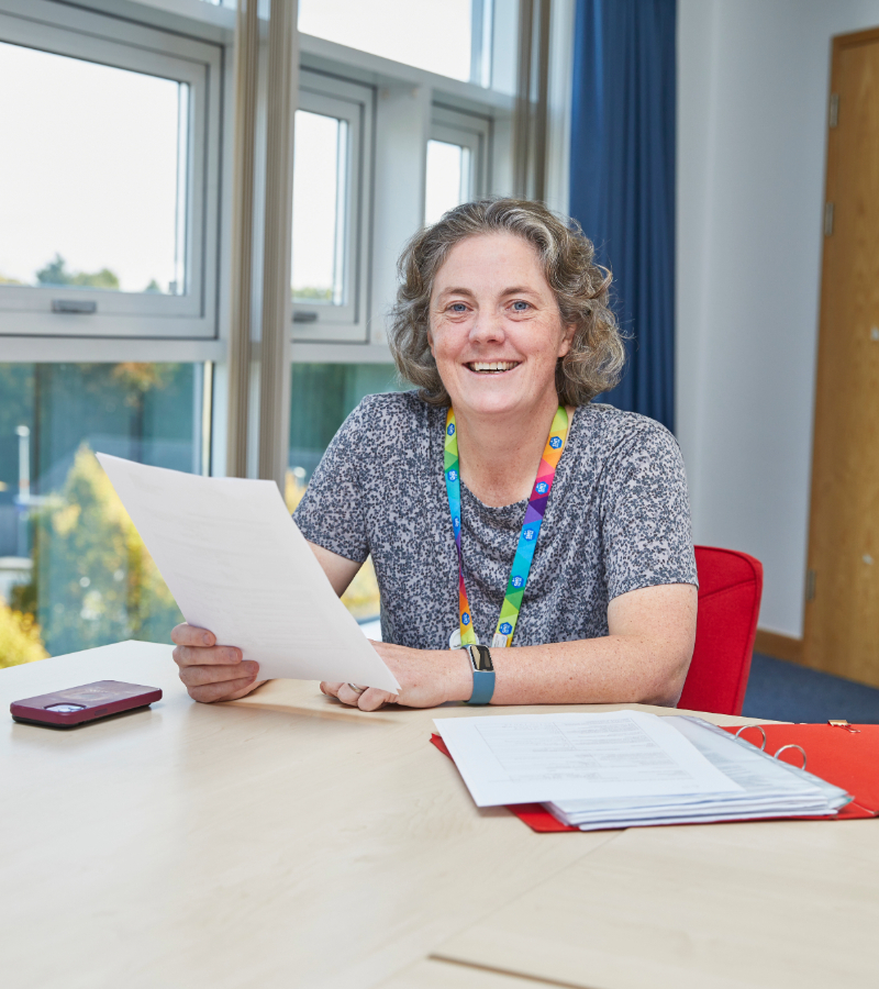 A woman with curly gray hair, smiling, seated at a desk holding documents in an office with large windows behind her. she wears a colorful lanyard and a casual outfit.