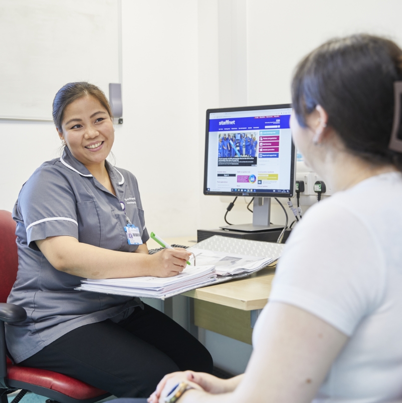 A smiling healthcare worker in scrubs sits at a desk with paperwork, facing a patient, with a computer screen displaying health-related information in the background.