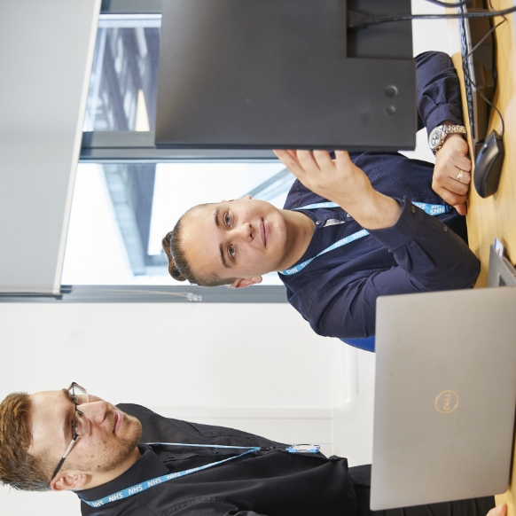 Two male professionals, one sitting at a desk with a laptop and the other standing beside him, both looking at a computer screen in a modern office setting.