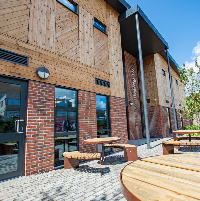 Exterior of a modern building with a mixture of wood and brick design, featuring large windows, a glass entrance, and circular wooden tables in the foreground.