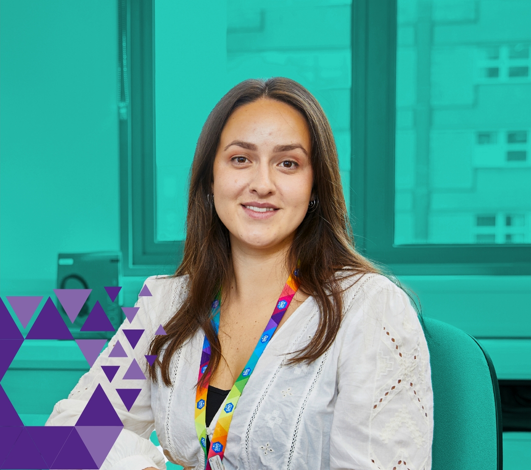 A professional woman with shoulder-length brown hair and a white blouse sits in an office. she wears a colourful lanyard and there's a green wall with windows in the background.