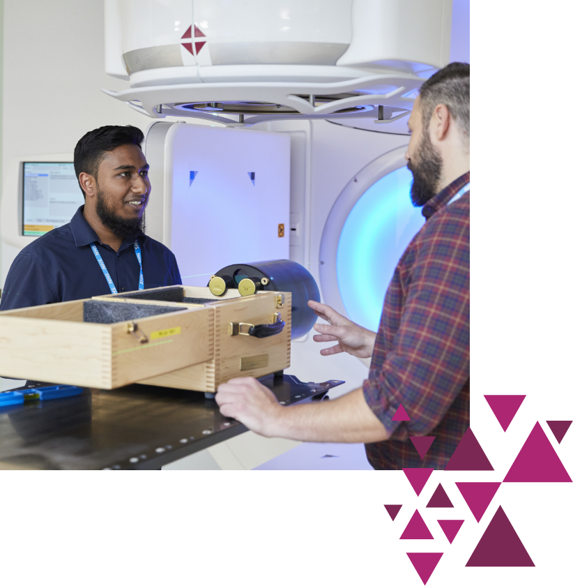 Two men, one in business casual and the other in a plaid shirt, standing and conversing in a hospital room with a large medical imaging machine in the background.