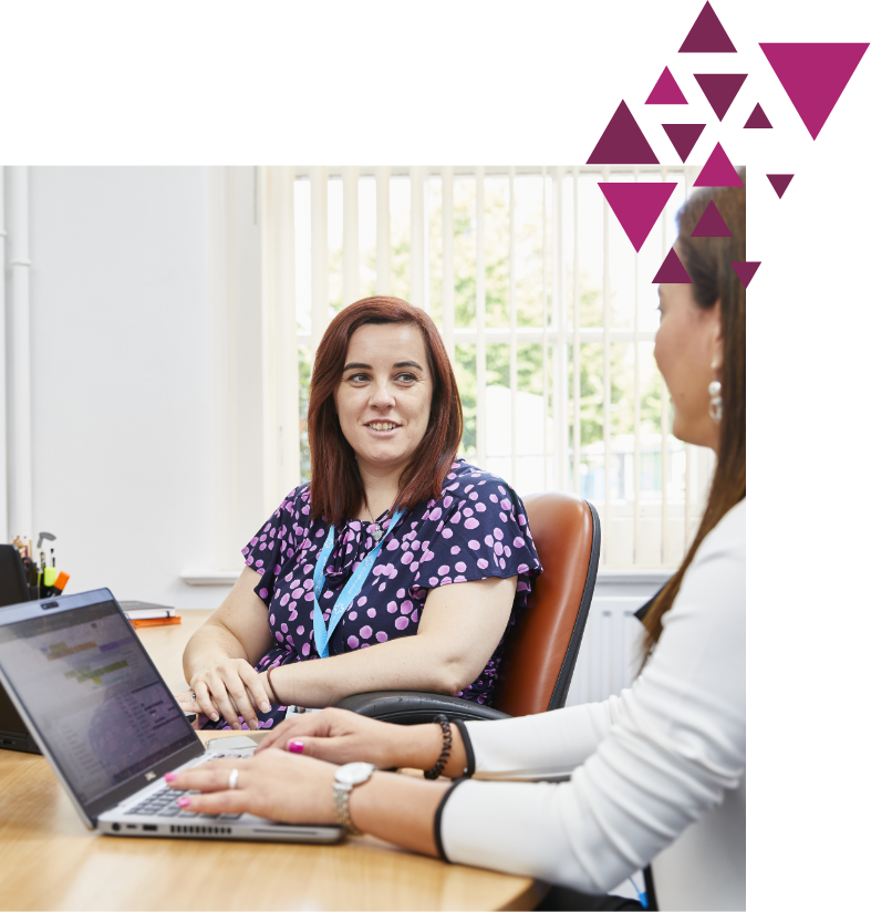 A woman in a purple patterned blouse smiles during a conversation with another woman at a desk, both using a laptop, in a bright office setting, decorated with purple geometric shapes.