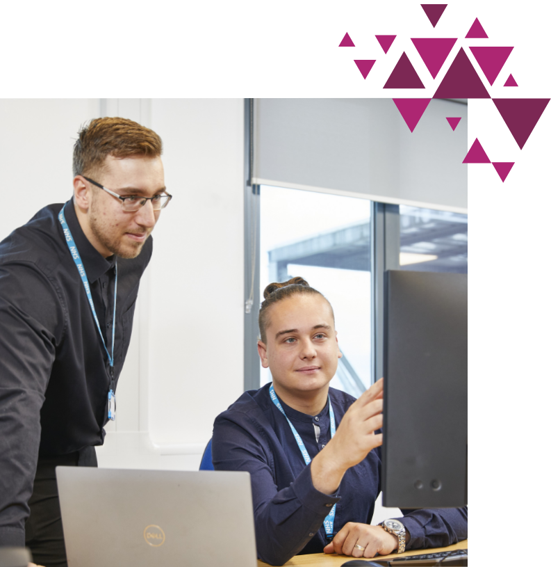 Two professionals, one standing, one seated, working together at a desk with a laptop and monitor. decorative purple geometric shapes are in the upper right corner. both are wearing id badges.