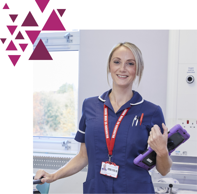 A smiling female nurse in a blue uniform holding a portable device, standing in a hospital room with a window showing autumn foliage. she has a lanyard and a badge.