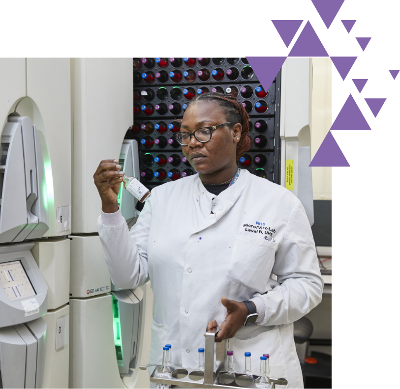 A female scientist in a white lab coat examines a vial in a laboratory filled with advanced equipment and colorful test tube racks in the background.