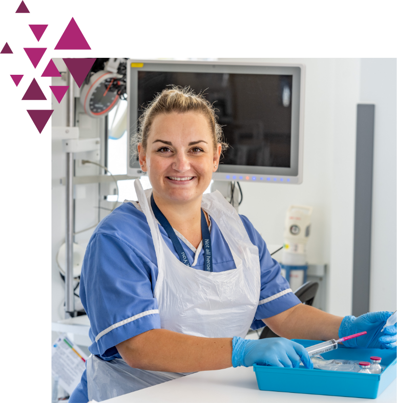 A female nurse in blue scrubs and gloves prepares medical instruments, smiling at the camera in a well-equipped hospital room.