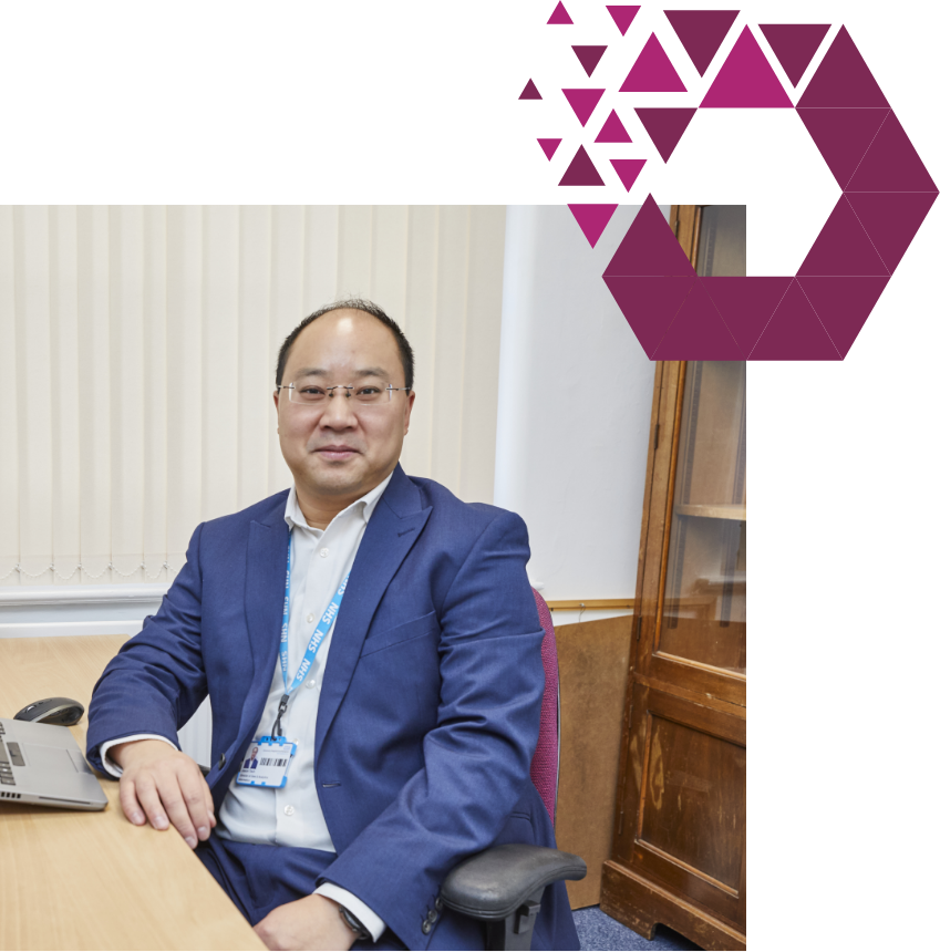 A man in a business attire with a lanyard, sitting at a desk in an office setting, smiles at the camera. a geometric purple shape decorates the top right corner.