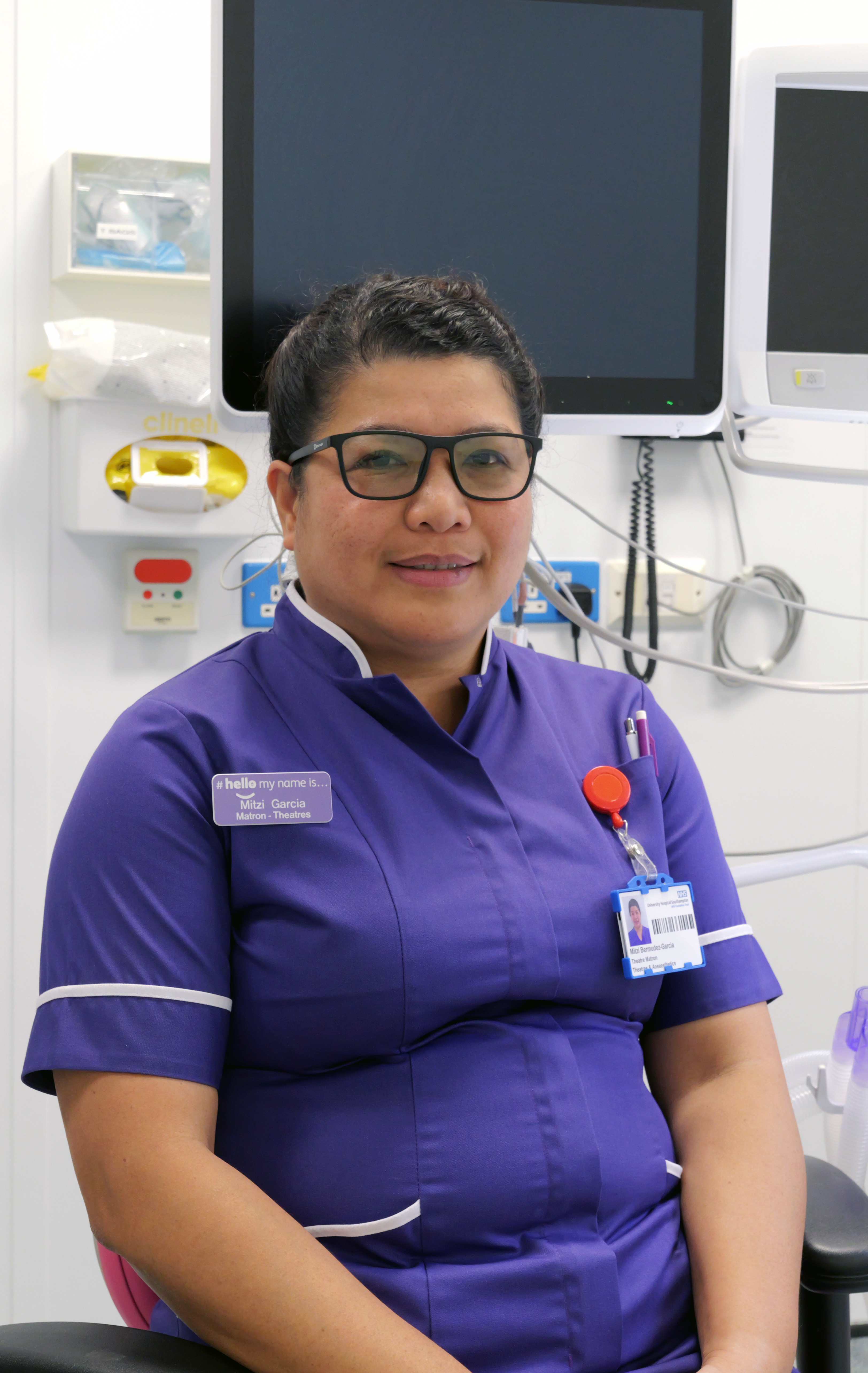 A smiling healthcare worker in a purple uniform, wearing glasses and a name tag, sitting in a clinical room with medical equipment in the background.