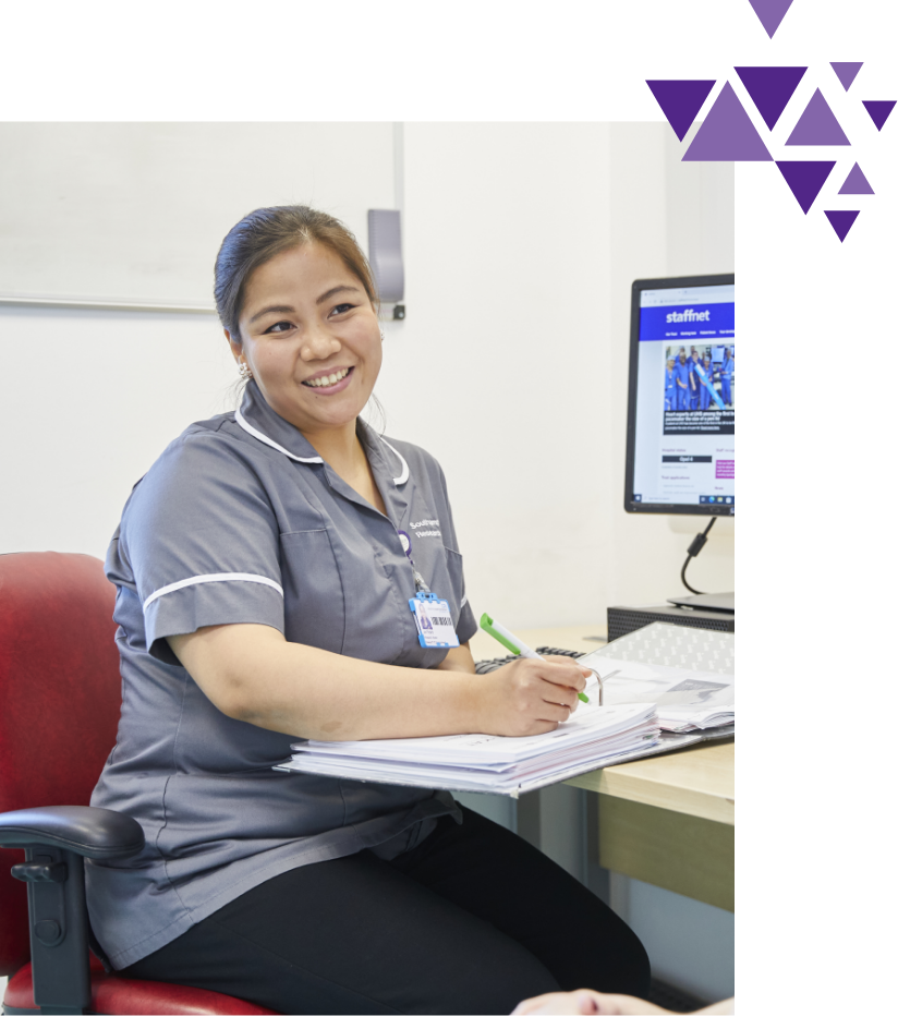 A smiling nurse in scrubs sitting at a desk, writing on documents beside a computer displaying a staffing program, with abstract purple shapes in the upper right corner.