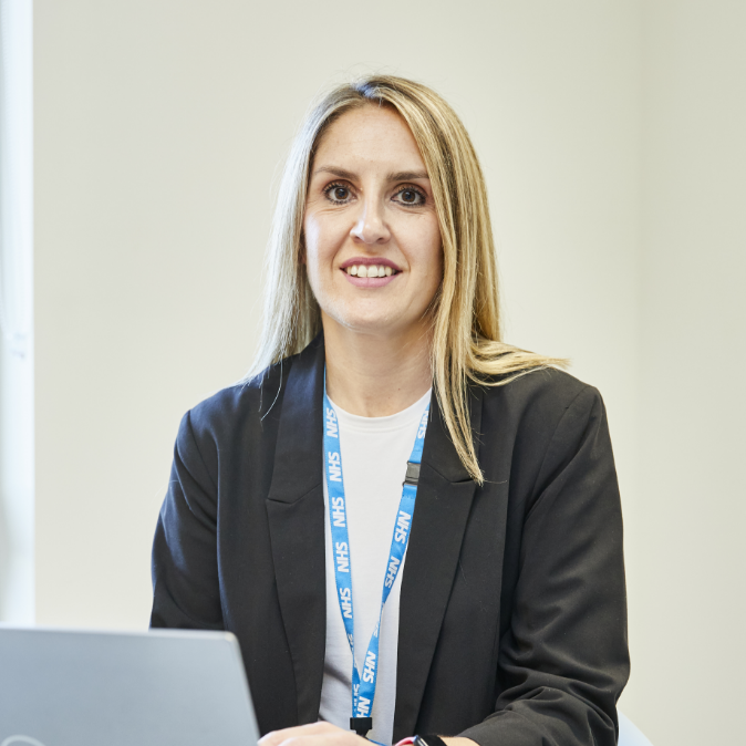 A professional woman with long blonde hair, wearing a dark blazer and an id badge, sits at a desk with a laptop, smiling at the camera in an office setting.