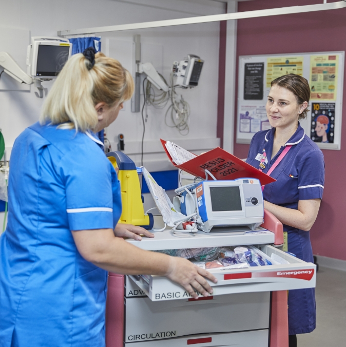 Two female medical professionals in a hospital, one in blue and one in navy scrubs, interact over a medical trolley stocked with equipment and supplies.